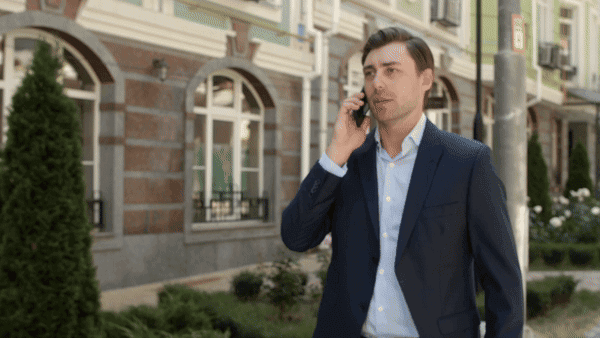 Businessman in a suit walking down a city street while talking on a smartphone, with a backdrop of elegant buildings and greenery.